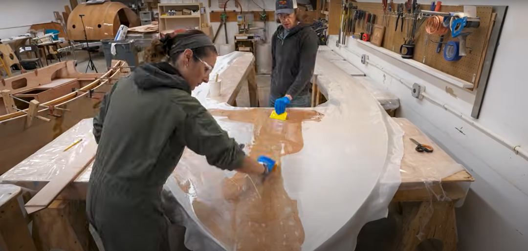 Sophie and Ryan have poured epoxy onto the foredeck and are scraping it smooth to completely cover the fiberglass cloth.