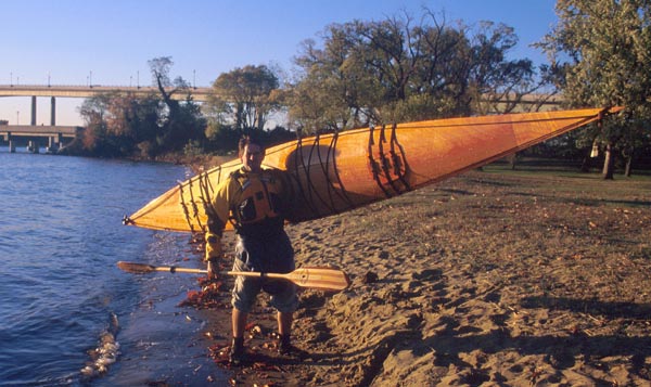 A young man carrying a Chesapeake 16 wooden kayak up onto a beach