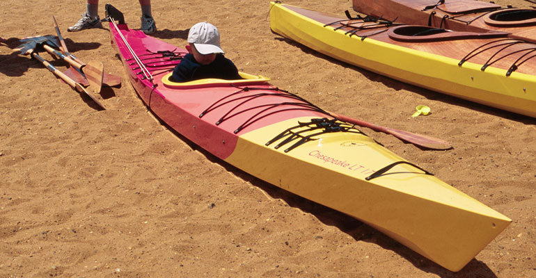 A youngster resting in the cockpit of a beached Chesapeake 17LT wooden kayak with a custom red and yellow paint job