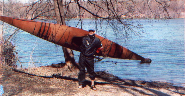 A man carrying a Chesapeake 17 wooden kayak on his shoulder at lakeside
