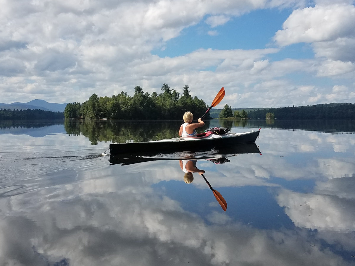 A woman paddling a Chesapeake 17 wooden kayak built by Ronald E. on the beautifully reflective Union Falls Pond in the Adirondacks