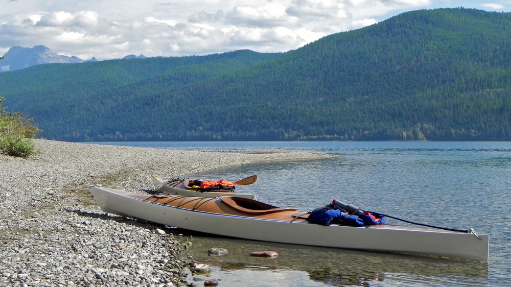 A Chesapeake 17 wooden kayak near a Chesapeake 16LT, both built by Harrison B., on a stony beach at McDonald Lake in Montana's Glacier National Park
