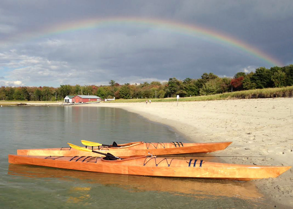 A pair of Chesapeake 17 wooden kayaks built by Mike D. under a rainbow on a beach in Portland, ME