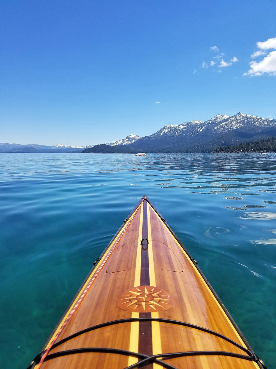The strip-built foredeck with inlaid compass rose of a Chesapeake 18 hybrid wooden kayak with a view of the mountains beyond Lake Tahoe
