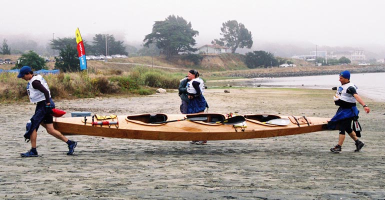 Three paddlers carrying a Chesapeake Triple wooden kayak ashore after a race. Built by Mark M.