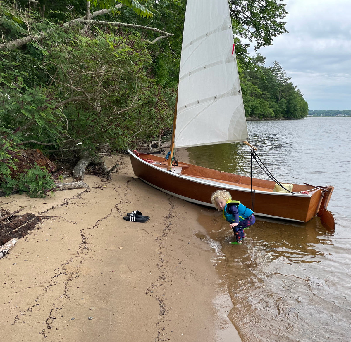 Jimmy Skiff on a beach with the sail up and a child playing in the water. 